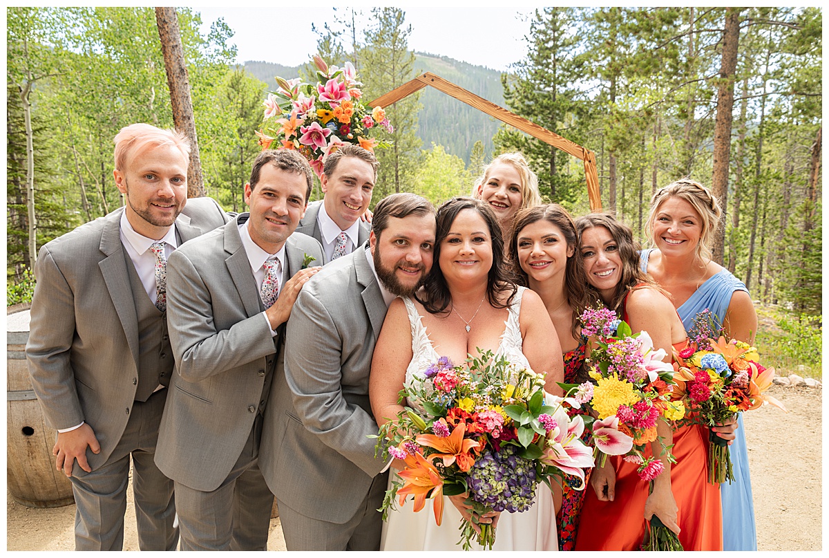 The bride, groom, and bridal party are posing for photos. The men are wearing gray suits and the women are in different colored dresses. Pine trees and a mountain are behind them.