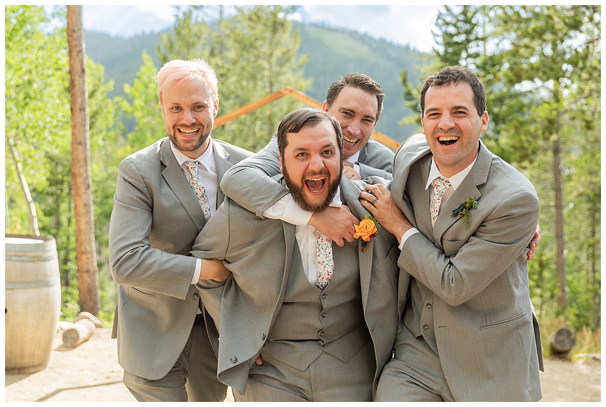 The groom and groomsmen are posing for photos. The men are wearing gray suits. Pine trees and a mountain are behind them.