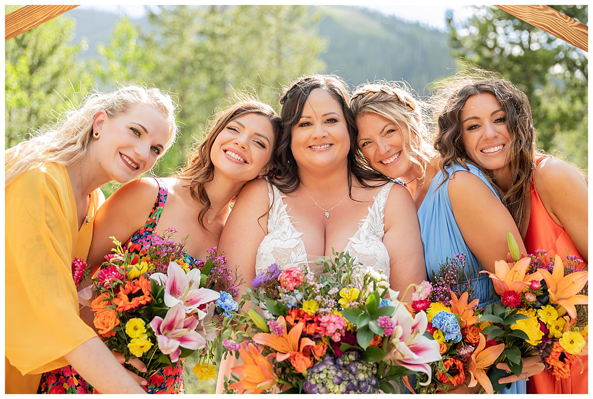 The bride and bridesmaids are posing for photos. The bridesmaids are all wearing different colored dresses. Pine trees and a mountain are behind them.