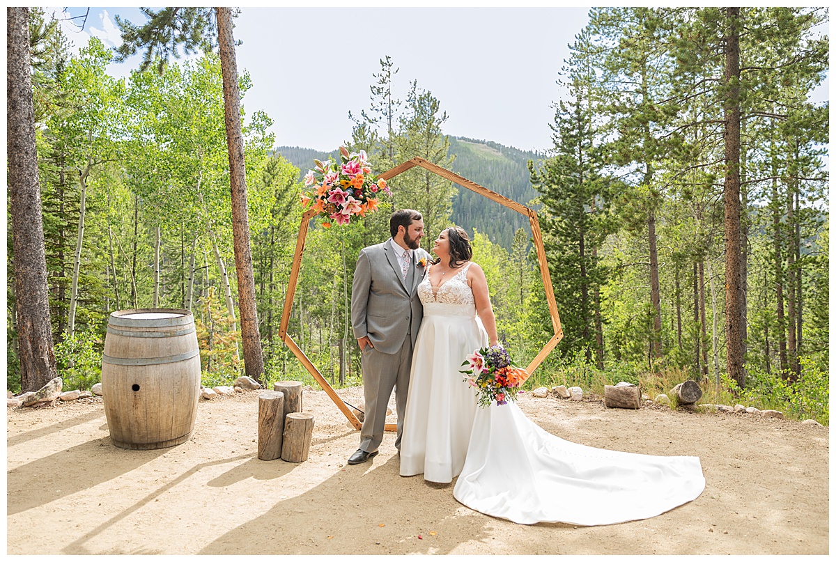 The bride and groom pose for photos. Their wedding arch, pine trees, and a mountain are behind them.