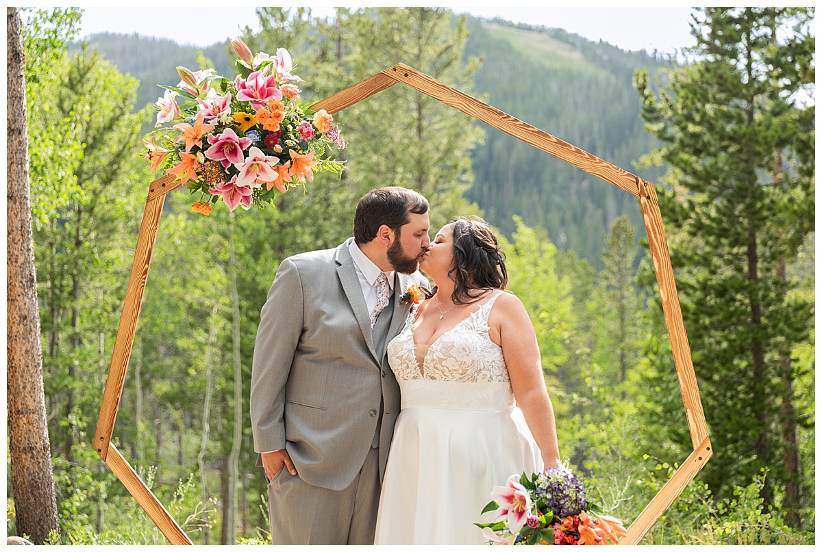 The bride and groom pose for photos. Their wedding arch, pine trees, and a mountain are behind them.