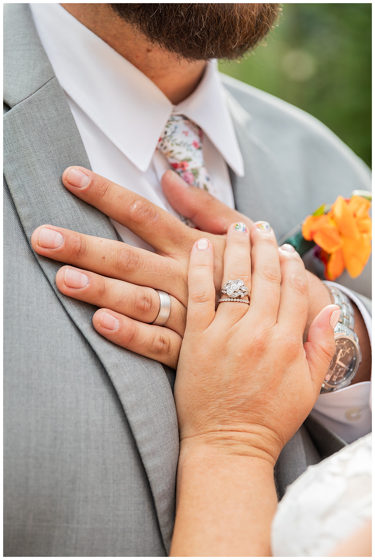 A close up photo of the couple's rings on their fingers. Their hands are placed on the groom's chest.