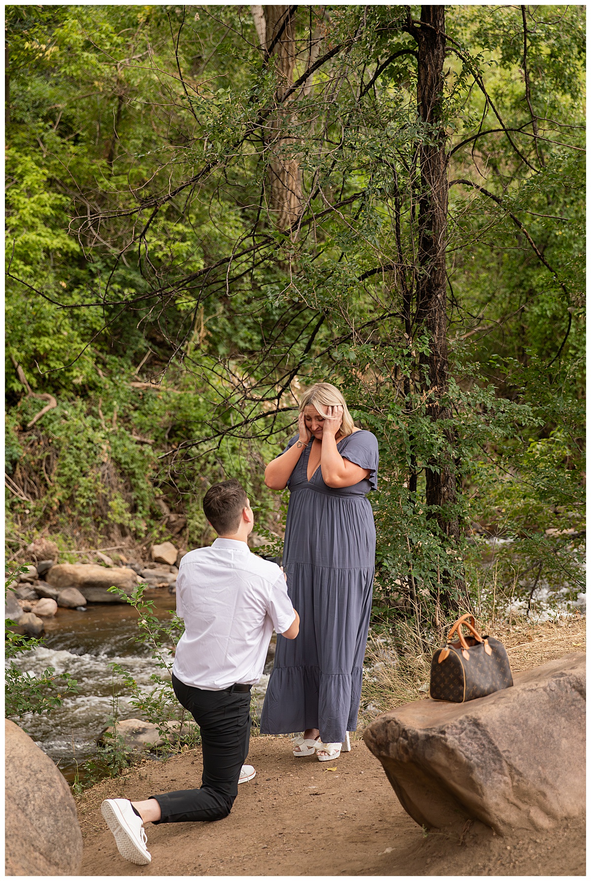 A man proposes to his girlfriend. He is on one knee wearing black slacks and a short sleeve, white button-down. She has short blonde hair and is wearing a long blue dress. There are green trees and a river in the background.