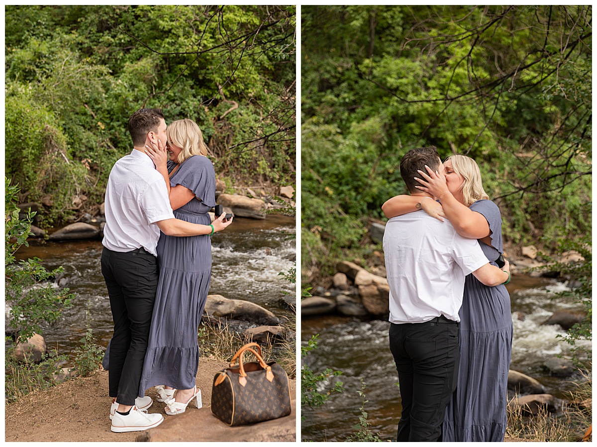 The couple celebrates after the proposal. He is wearing black slacks and a short sleeve, white button-down. She has short blonde hair and is wearing a long blue dress. There are green trees and a river in the background.