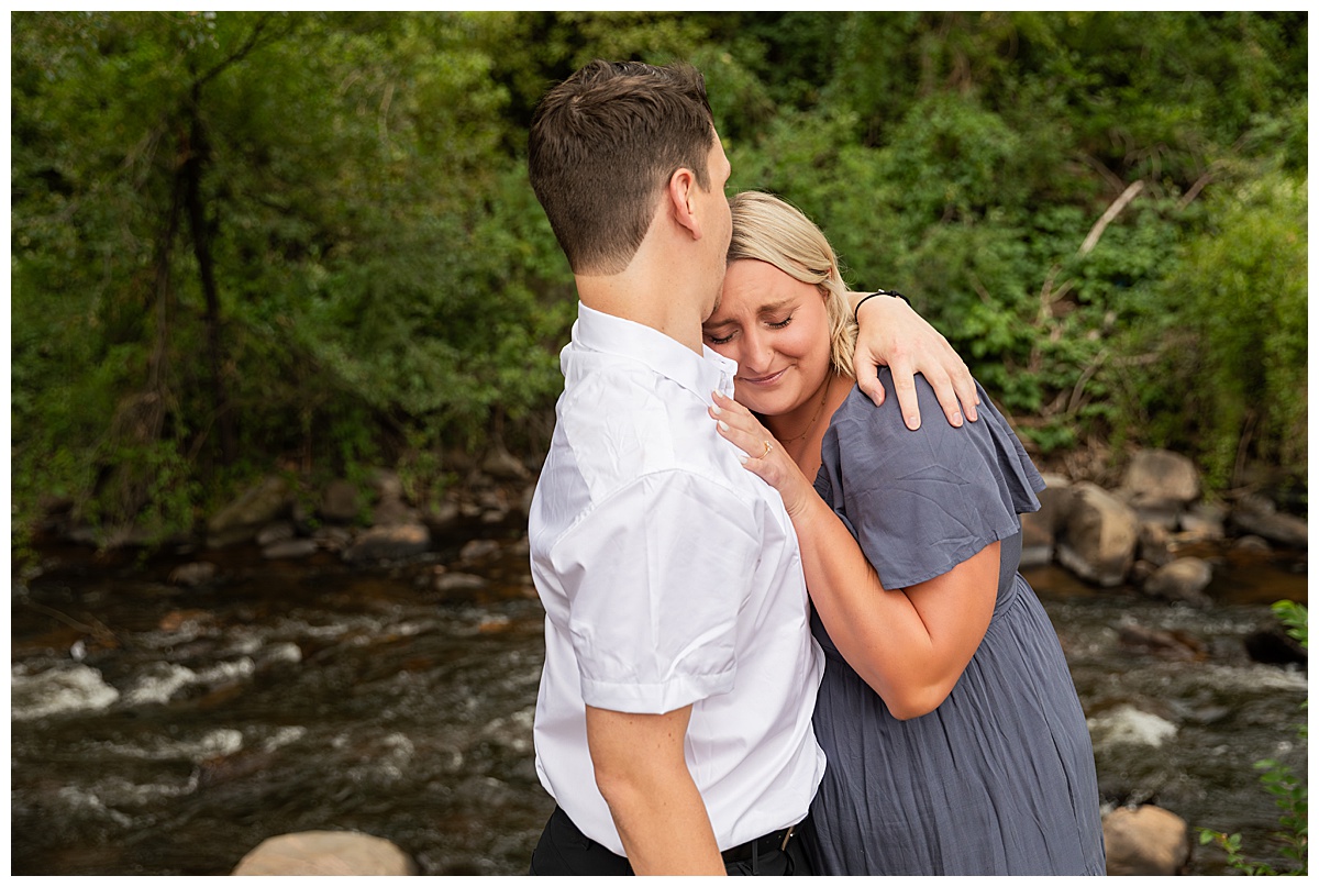 The couple celebrates after the proposal. He is wearing black slacks and a short sleeve, white button-down. She has short blonde hair and is wearing a long blue dress. There are green trees and a river in the background.