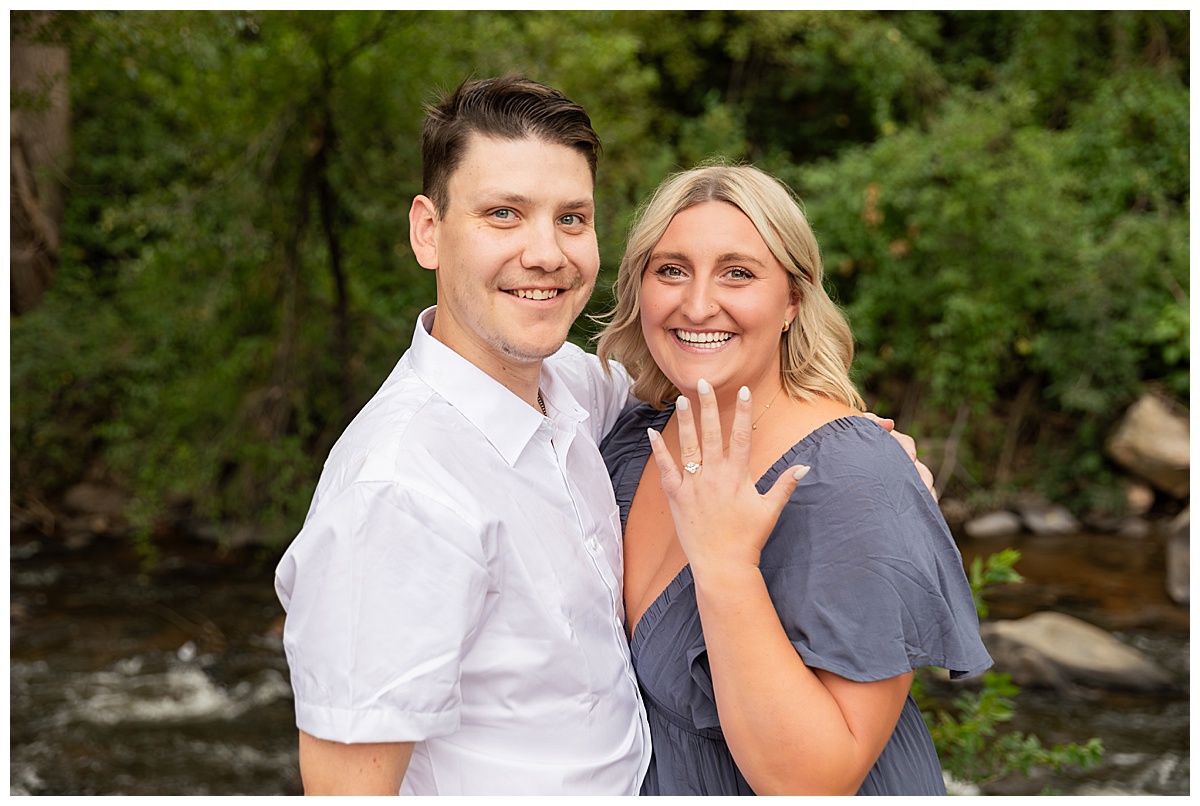 The couple celebrates after the proposal. He is wearing black slacks and a short sleeve, white button-down. She has short blonde hair and is wearing a long blue dress. There are green trees and a river in the background.