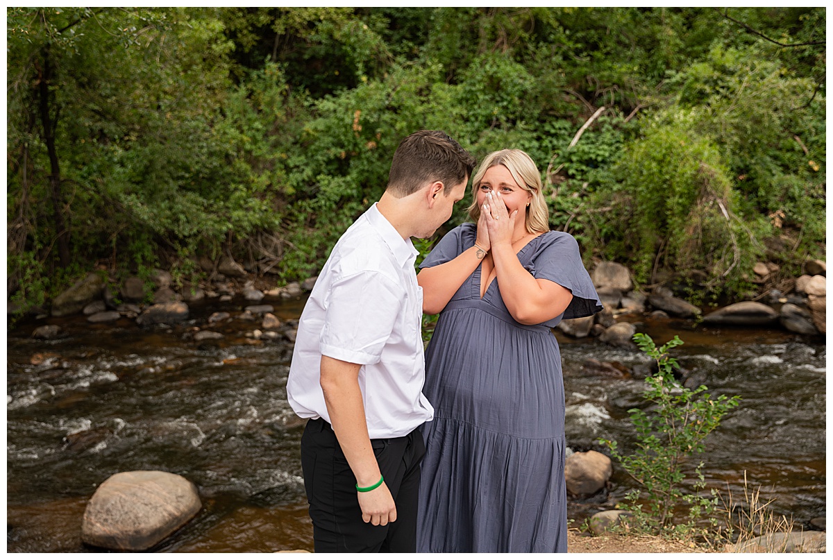 The couple celebrates after the proposal. He is wearing black slacks and a short sleeve, white button-down. She has short blonde hair and is wearing a long blue dress. There are green trees and a river in the background.