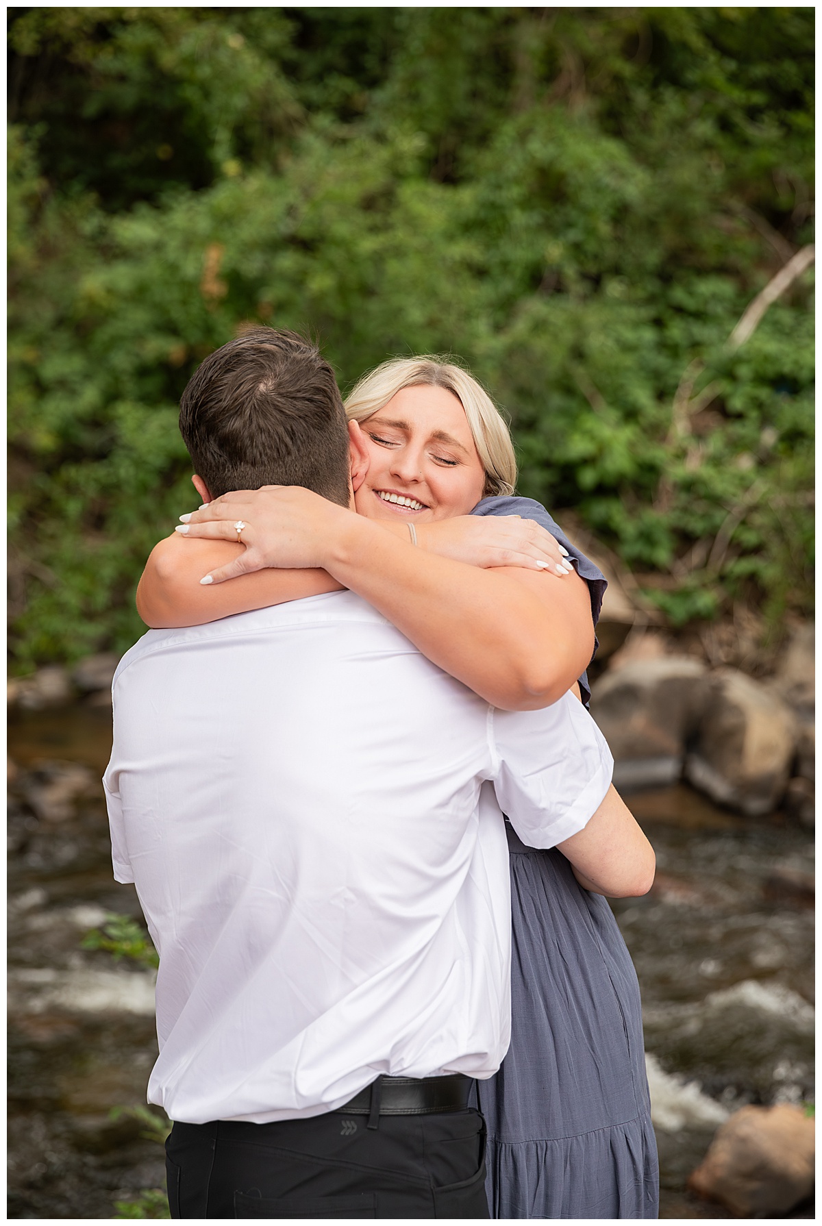 The couple celebrates after the proposal. He is wearing black slacks and a short sleeve, white button-down. She has short blonde hair and is wearing a long blue dress. There are green trees and a river in the background.