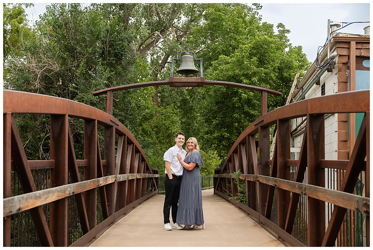 The couple celebrates after the proposal. He is wearing black slacks and a short sleeve, white button-down. She has short blonde hair and is wearing a long blue dress. They are standing on a bridge and there are green trees and a river in the background.