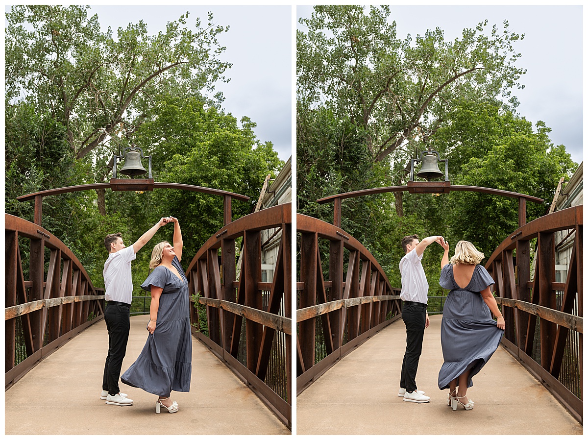 The couple celebrates after the proposal. He is wearing black slacks and a short sleeve, white button-down. She has short blonde hair and is wearing a long blue dress. They are standing on a bridge and there are green trees and a river in the background.