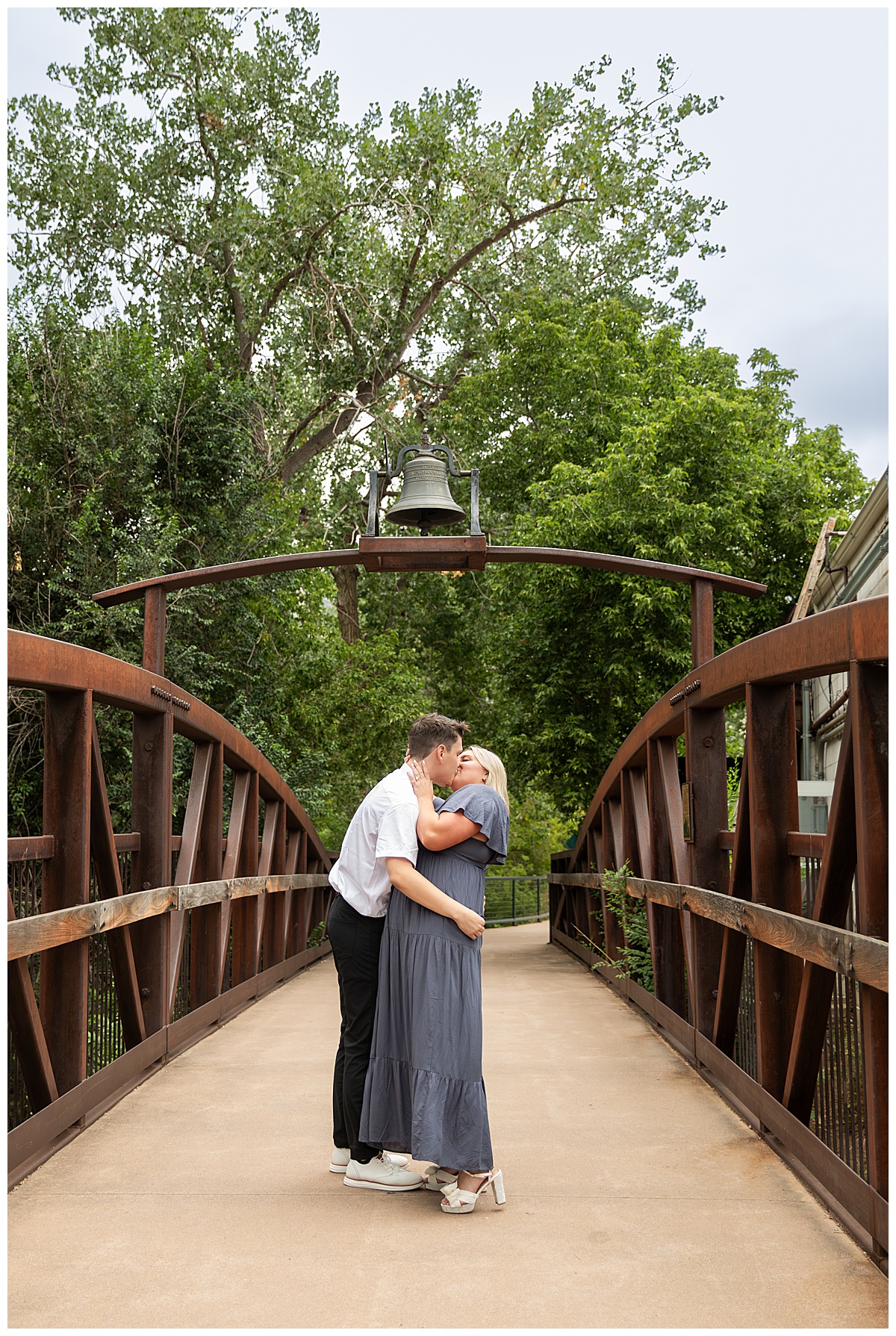 The couple celebrates after the proposal. He is wearing black slacks and a short sleeve, white button-down. She has short blonde hair and is wearing a long blue dress. They are standing on a bridge and there are green trees and a river in the background.