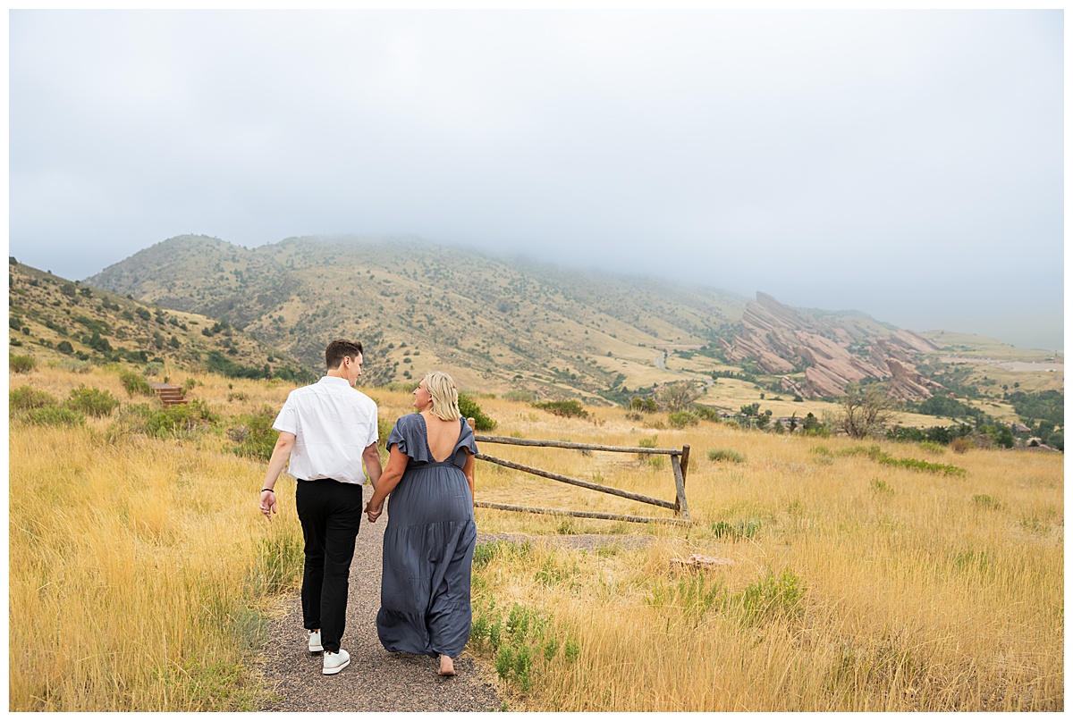 The couple takes couple's portraits. He is wearing black slacks and a short sleeve, white button-down. She has short blonde hair and is wearing a long blue dress. In a field in front of Red Rocks Amphitheater. 