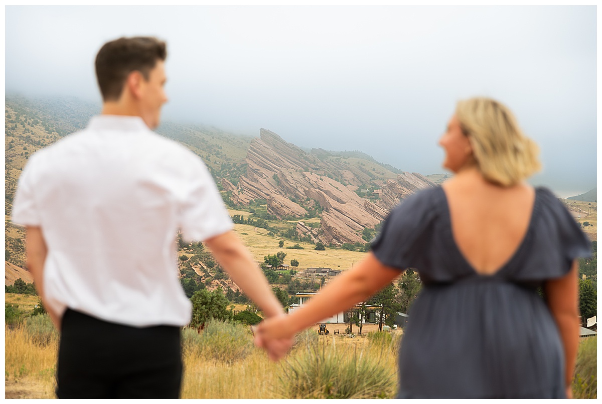 The couple takes couple's portraits. He is wearing black slacks and a short sleeve, white button-down. She has short blonde hair and is wearing a long blue dress. In a field in front of Red Rocks Amphitheater. 