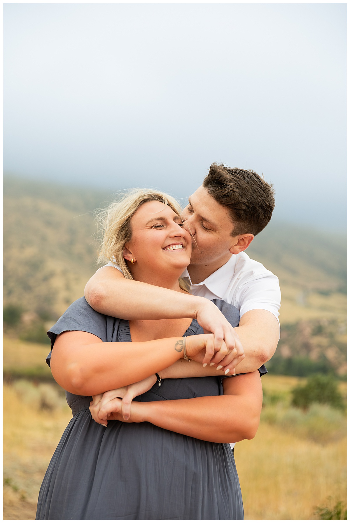 The couple takes couple's portraits. He is wearing black slacks and a short sleeve, white button-down. She has short blonde hair and is wearing a long blue dress. In a field in front of Red Rocks Amphitheater. 