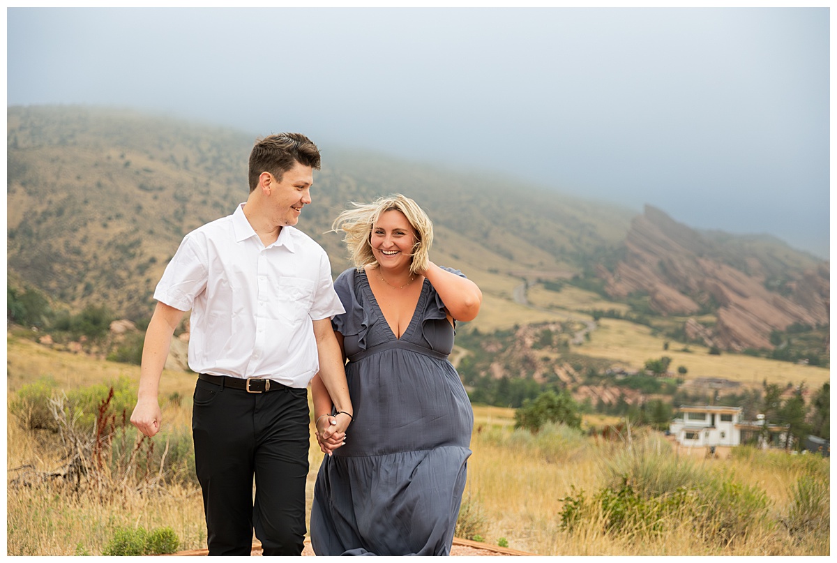 The couple takes couple's portraits. He is wearing black slacks and a short sleeve, white button-down. She has short blonde hair and is wearing a long blue dress. In a field in front of Red Rocks Amphitheater. 