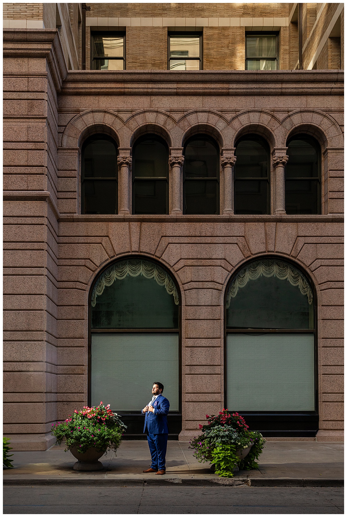 The groom gets ready in the hotel lobby. He has dark brown hair and a beard. He is wearing a blue suit with a floral tie.