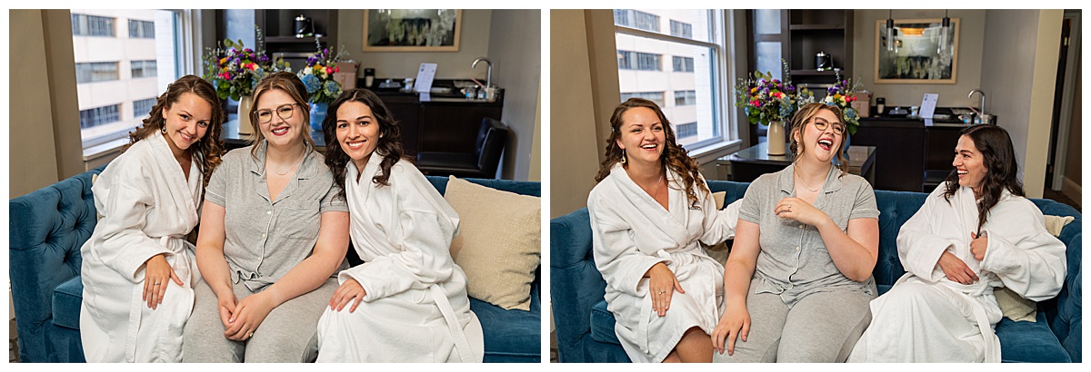 The bride gets ready with her two bridesmaids. She has brown hair in an updo and glasses. She is wearing a gray PJ set and the two bridesmaids are wearing white bathrobes. They are sitting on a blue couch.