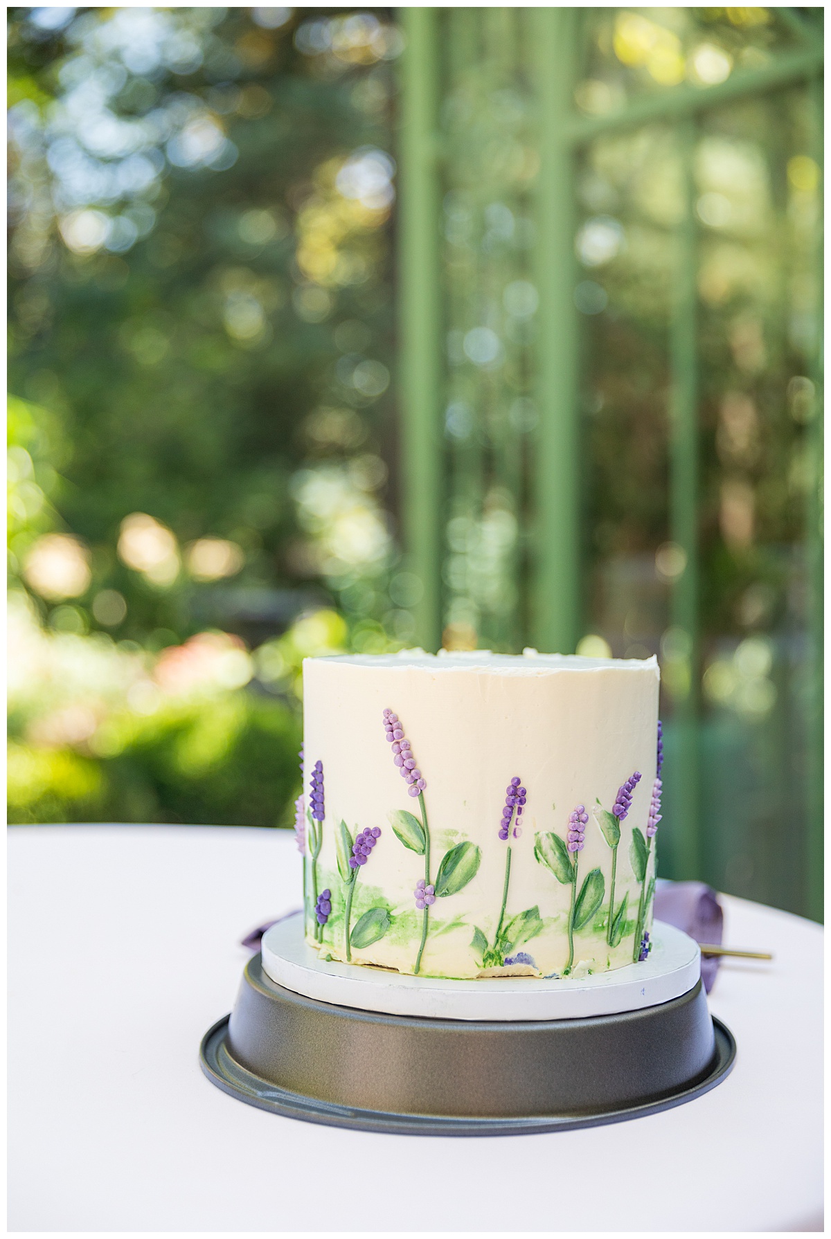 A close up of the cake outside of the green metal Woodland Mosaic Solarium. The cake is small with white icing; it has purple icing flowers with green stems and leaves on it.