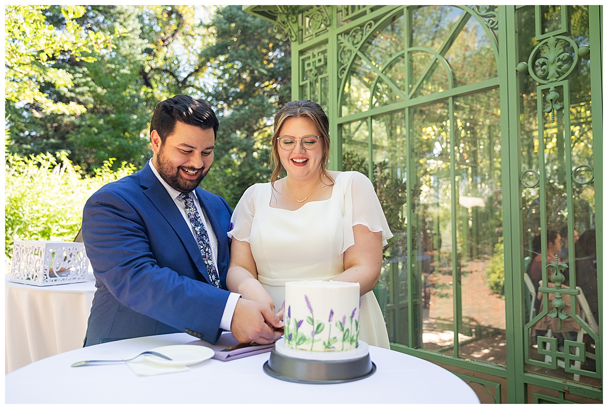 The bride and groom cut the cake outside of the green metal Woodland Mosaic Solarium. The cake is small with white icing; it has purple icing flowers with green stems and leaves on it.