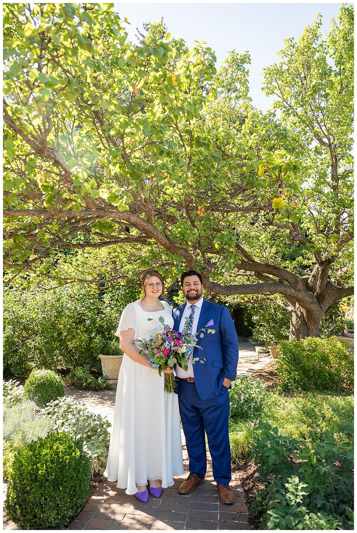 The bride and groom pose under a tree at their Denver Botanic Gardens wedding. They are smiling at the camera.