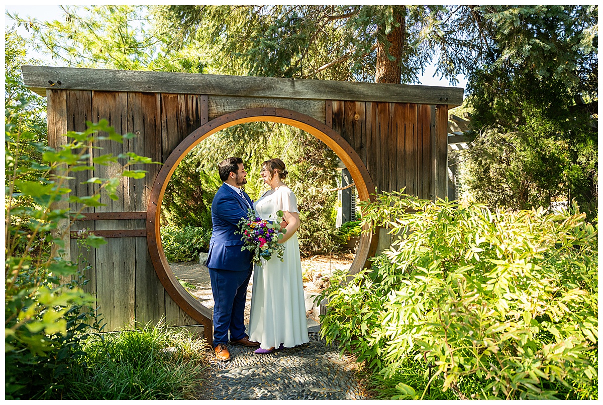 The bride and groom pose in the Asian garden. They are standing under a large wooden circle arch.