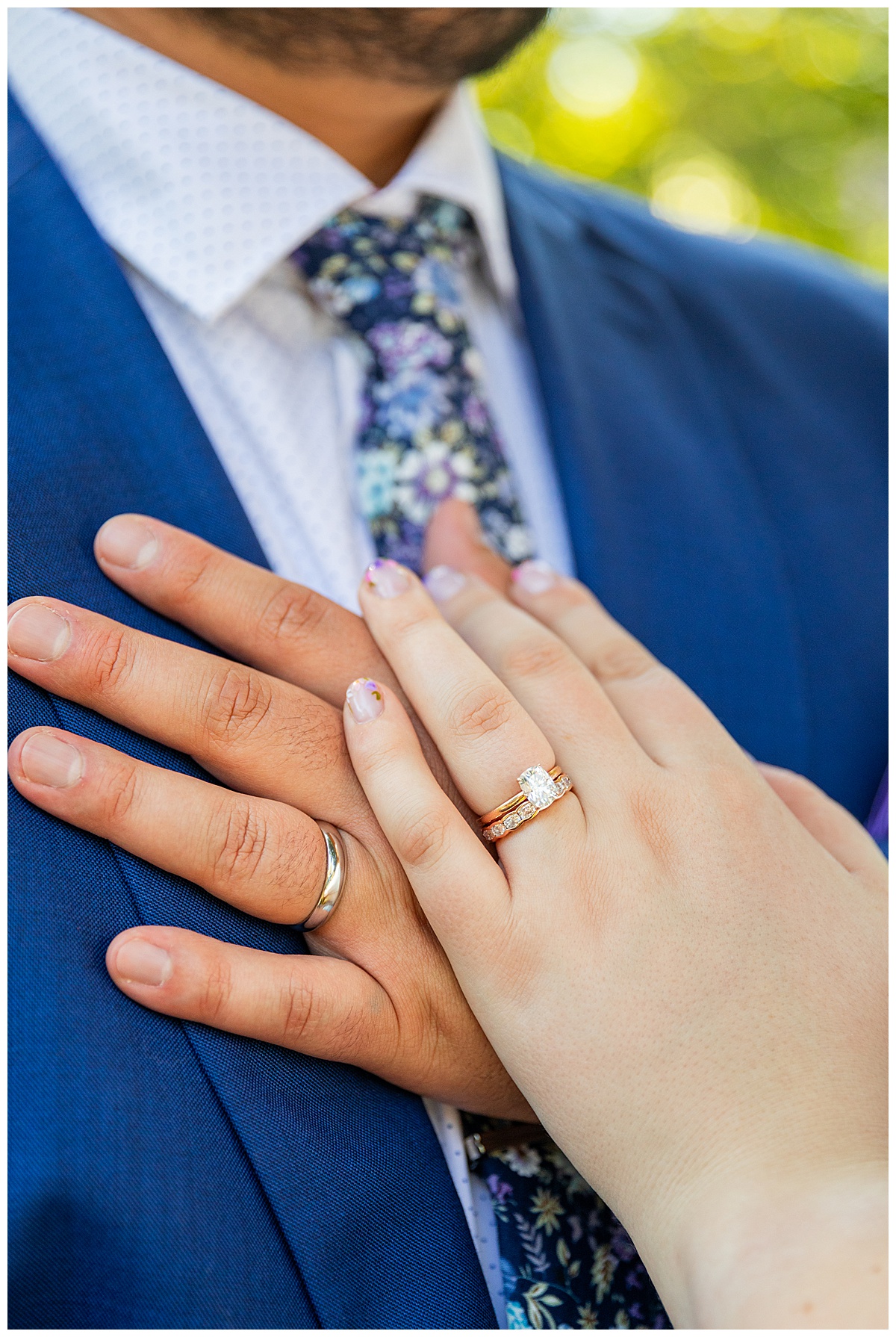 A close up of the rings on the couple's fingers. Their hands are resting on the groom's chest.