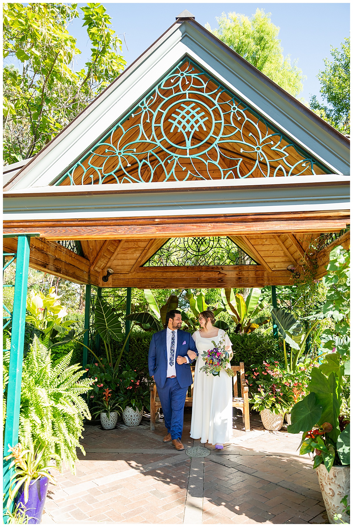 The bride and groom walk with arms linked under the gazebo in the Victoria Secret Garden. The gazebo with wood with teal metal accents.