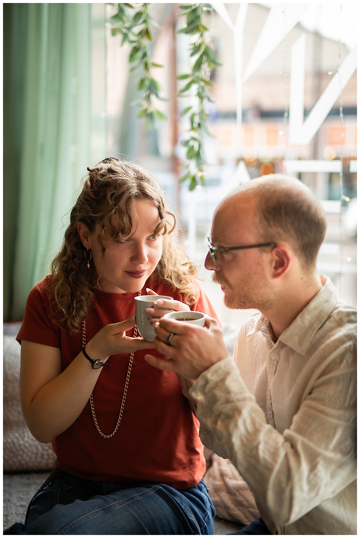 Lily & Cody enjoy tea sitting on a couch by a window. Lily has curly brown hair and is wearing a red shirt and jeans. Cody is wearing glasses with a cream button down shirt and jeans.