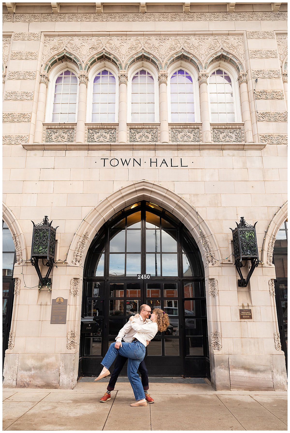 Lily & Cody pose in front of Downtown Littleton's Town Hall Art Center. Lily has curly brown hair and is wearing a red shirt, a cream jacket, and jeans. Cody is wearing glasses with a cream button down shirt and jeans.