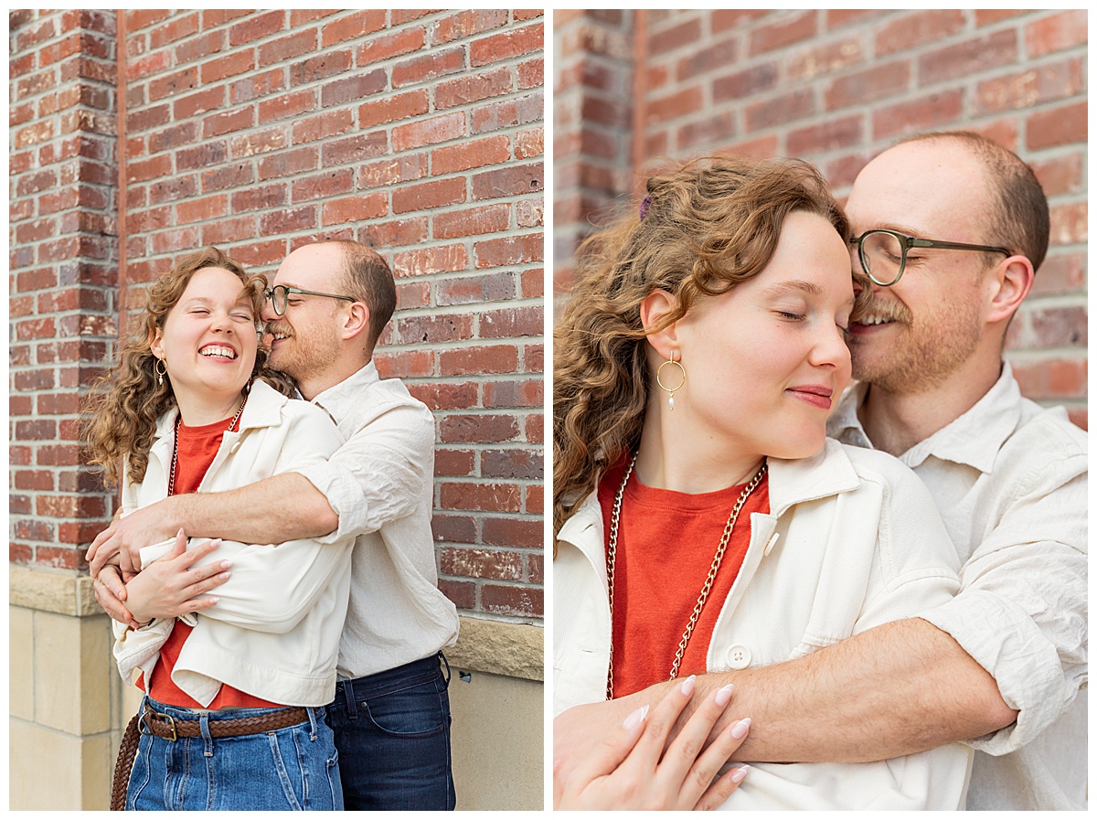 Lily & Cody pose in front of a red brick wall during their Downtown Littleton engagement session. He is cuddling her from behind. Lily has curly brown hair and is wearing a red shirt, a cream jacket, and jeans. Cody is wearing glasses with a cream button down shirt and jeans.