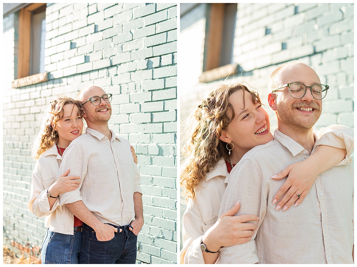 Lily & Cody pose in front of a blue brick wall during their Downtown Littleton engagement session. Lily has curly brown hair and is wearing a red shirt, a cream jacket, and jeans. Cody is wearing glasses with a cream button down shirt and jeans.