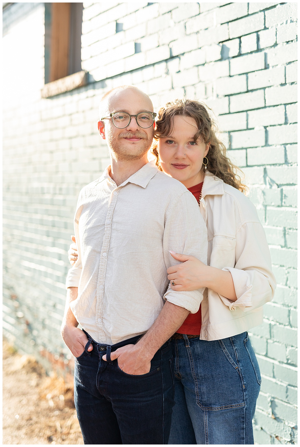 Lily & Cody pose in front of a blue brick wall during their Downtown Littleton engagement session. Lily has curly brown hair and is wearing a red shirt, a cream jacket, and jeans. Cody is wearing glasses with a cream button down shirt and jeans.