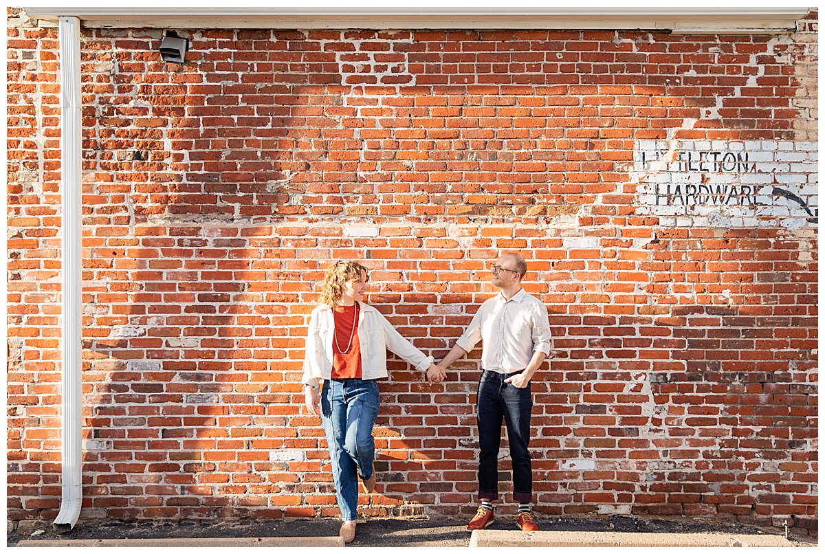 Lily & Cody pose in front of a red brick wall. Lily has curly brown hair and is wearing a red shirt, a cream jacket, and jeans. Cody is wearing glasses with a cream button down shirt and jeans.