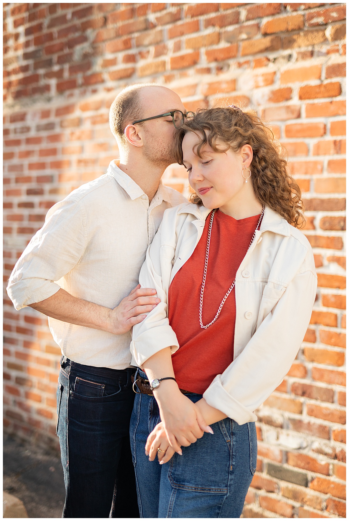 Lily & Cody pose in front of a red brick wall. Lily has curly brown hair and is wearing a red shirt, a cream jacket, and jeans. Cody is wearing glasses with a cream button down shirt and jeans.