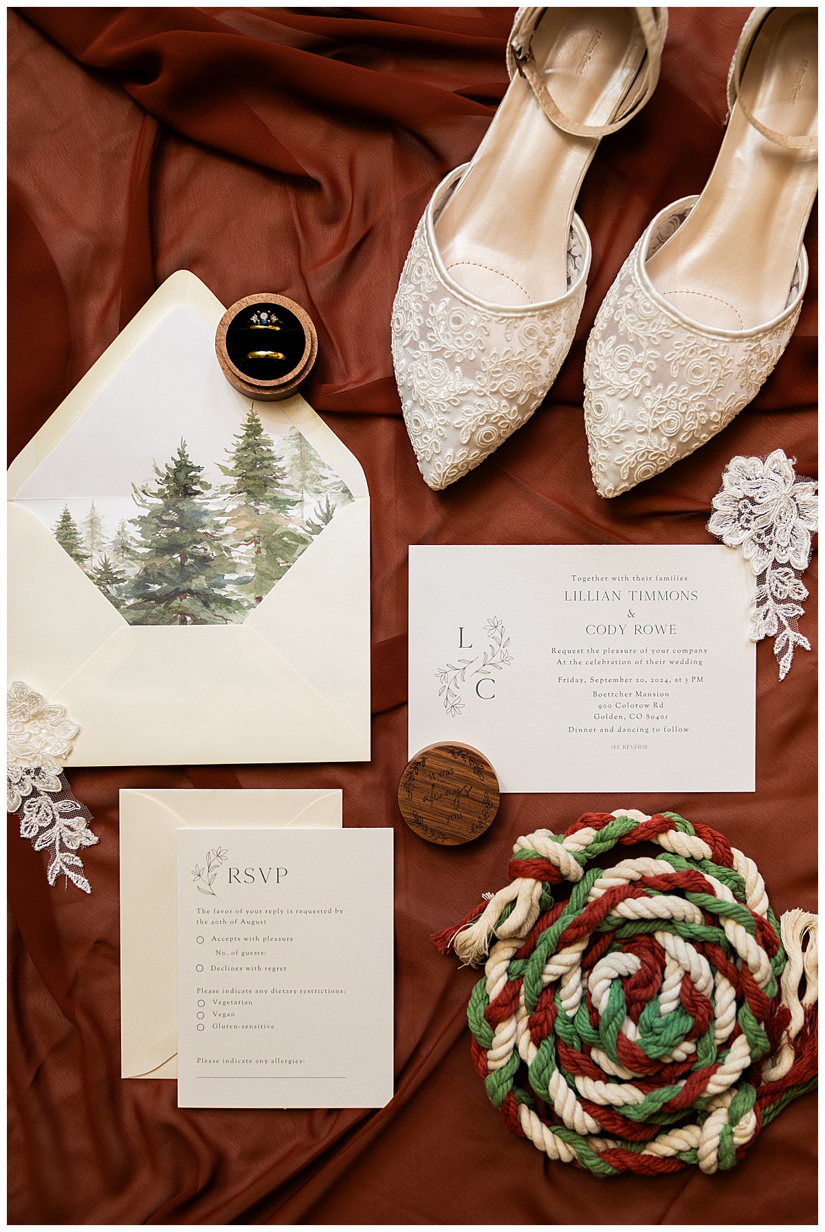 A close up of the couples details sitting on a burnt orange background. White lace shoes, scraps of lace flowers.,their wedding suite, and their wooden ring box. There is also their white, green, and red braided rope for their handfasting.