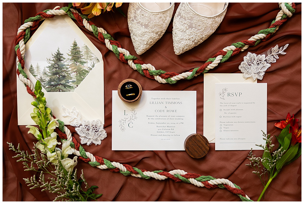 A close up of the couples details sitting on a burnt orange background. White lace shoes, scraps of lace flowers.,their wedding suite, and their wooden ring box. There is also their white, green, and red braided rope for their handfasting.