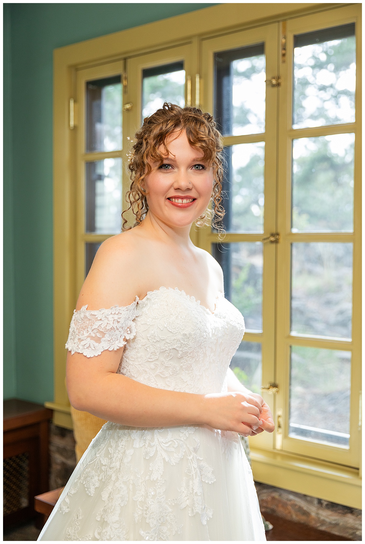 Photo of the bride after putting her dress on. She has curly dirty brown hair pulled up in an updo. Her dress is a lace a-line with a sweetheart neckline and off the shoulder straps.
