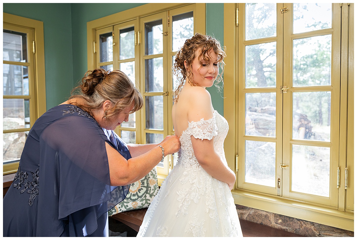 Photo of the bride's mom in her blue dress helping the bride into her wedding dress. She has curly dirty brown hair pulled up in an updo. Her dress is a lace a-line with a sweetheart neckline and off the shoulder straps.