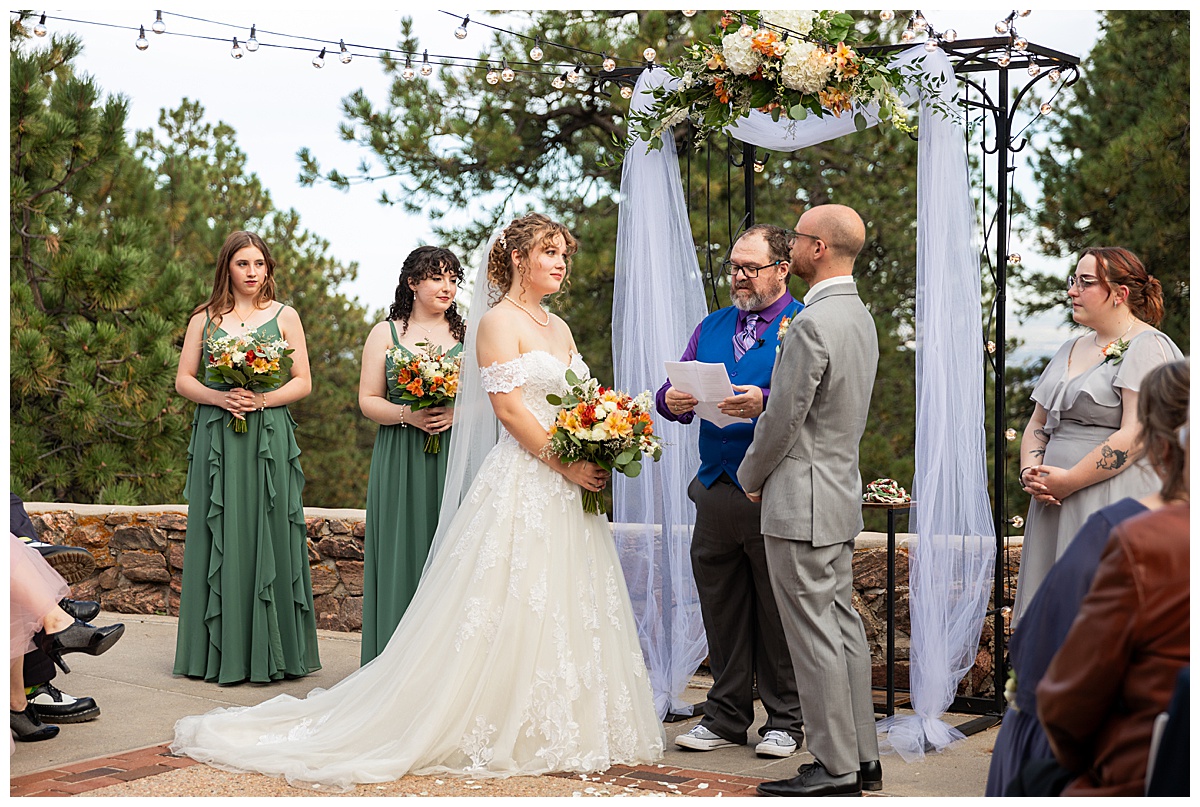 Photos of the bride and groom during their wedding ceremony. They are standing in front of a metal arch with white fabric draped over it and fall-colored flowers.