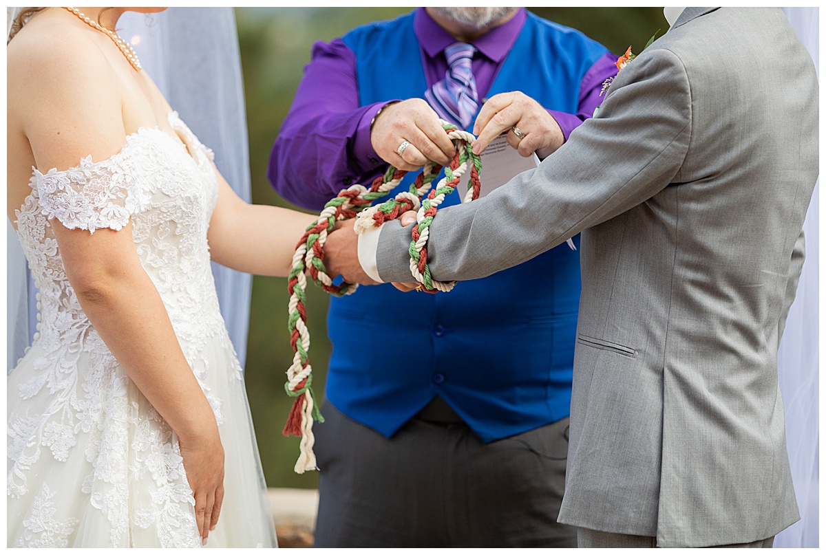 Photos of the bride and groom during their wedding ceremony. They are standing in front of a metal arch with white fabric draped over it and fall-colored flowers. They complete a traditional hand-fasting ceremony.
