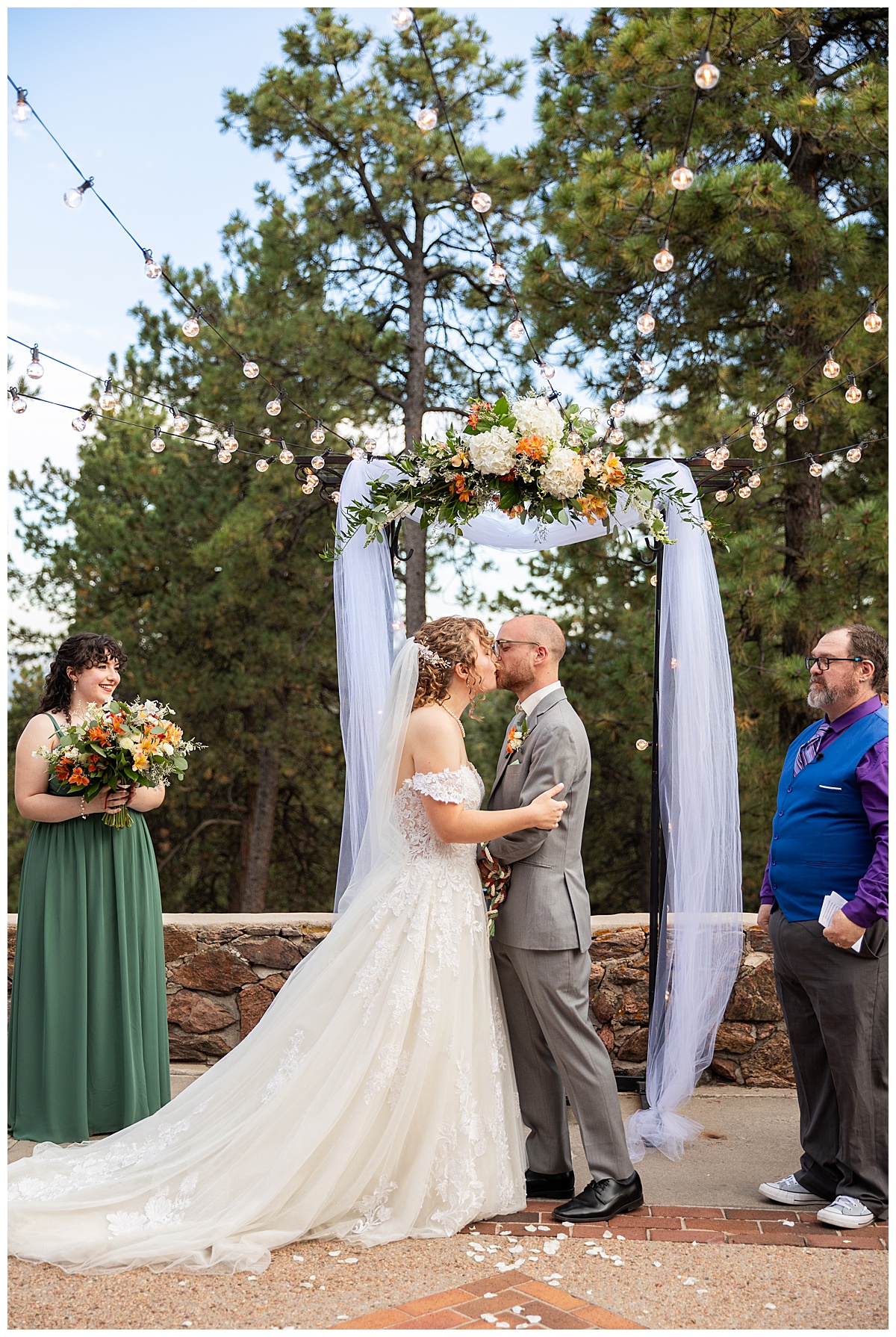 Photos of the bride and groom sharing their first kiss. They are standing in front of a metal arch with white fabric draped over it and fall-colored flowers.